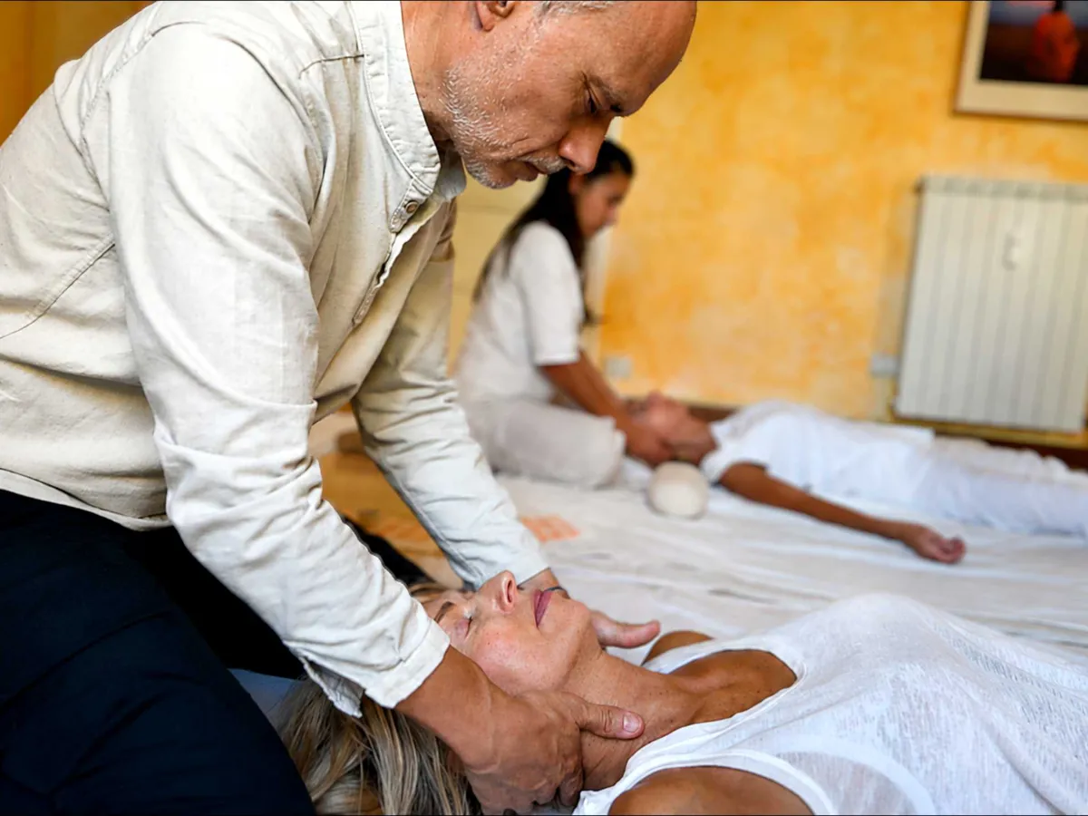 Un operatore Shiatsu sostiene con le mani il collo di una donna distesa su un futon durante un trattamento, mentre sullo sfondo un'altra coppia pratica un trattamento analogo nella sala della scuola